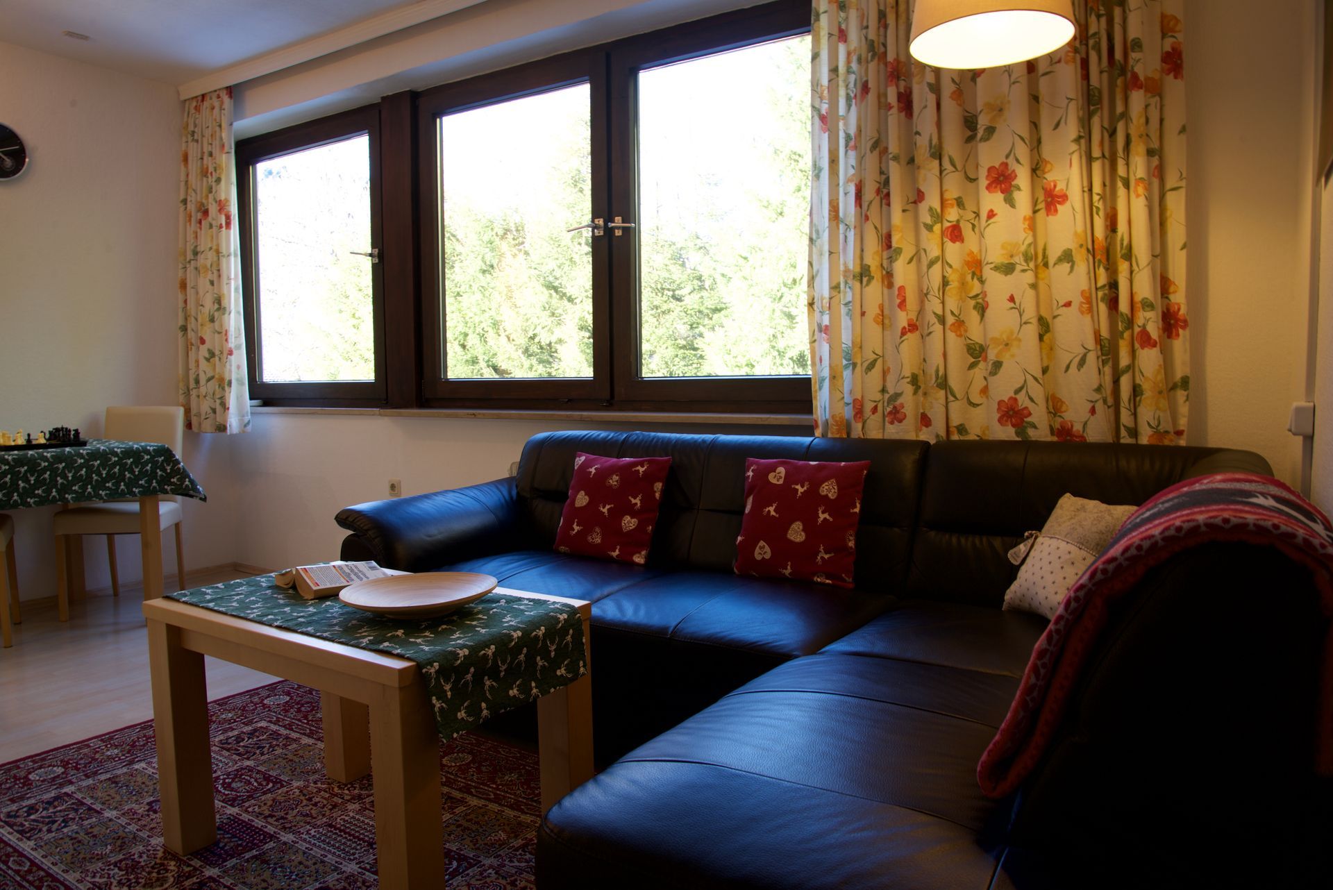 Gemütliches Wohnzimmer mit dunkelblauem Ecksofa, Holztisch, großen Fenstern und geblümten Vorhängen