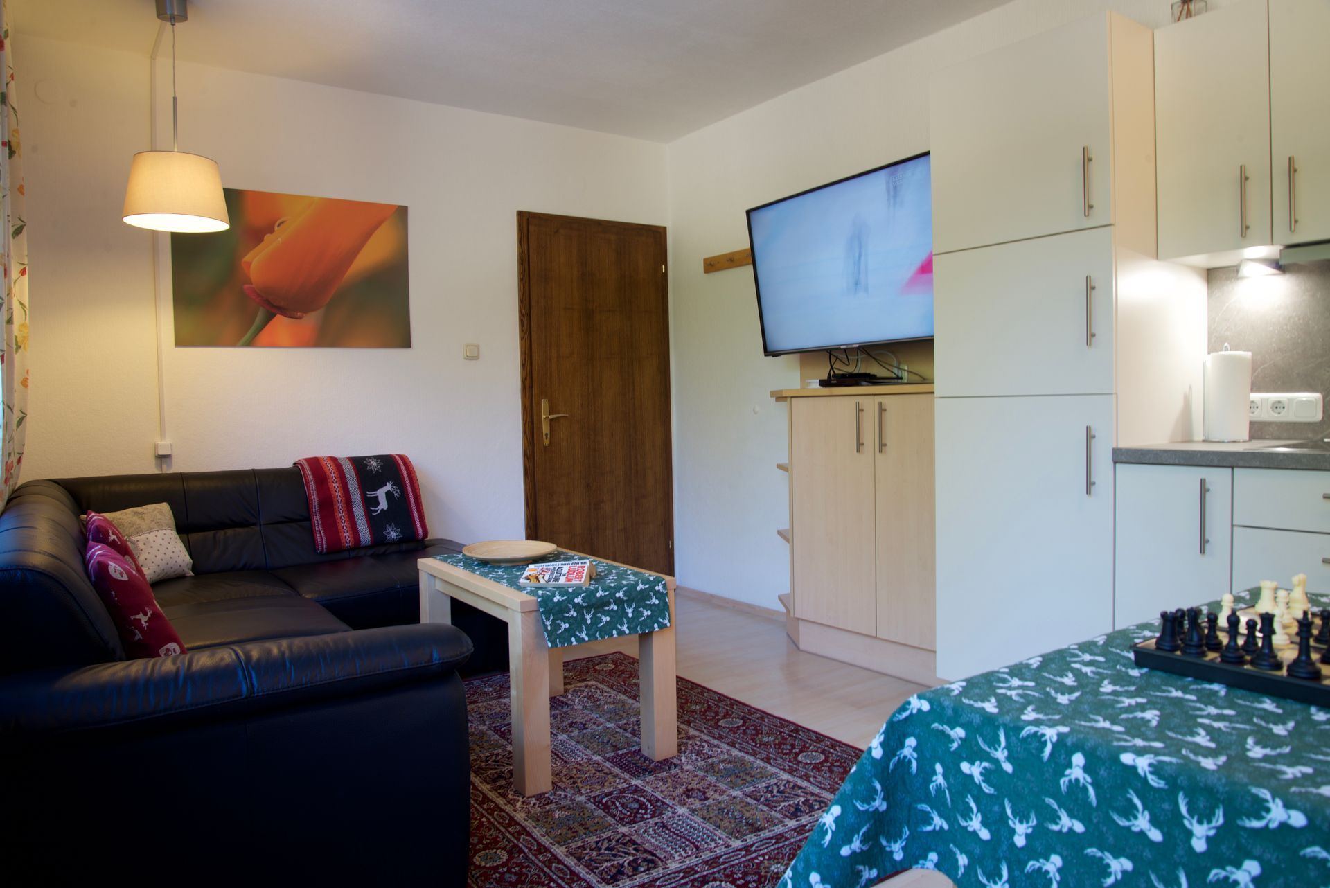 Kleines Wohnzimmer mit schwarzem Sofa, Couchtisch, an der Wand montiertem Fernseher und angrenzendem Küchenbereich