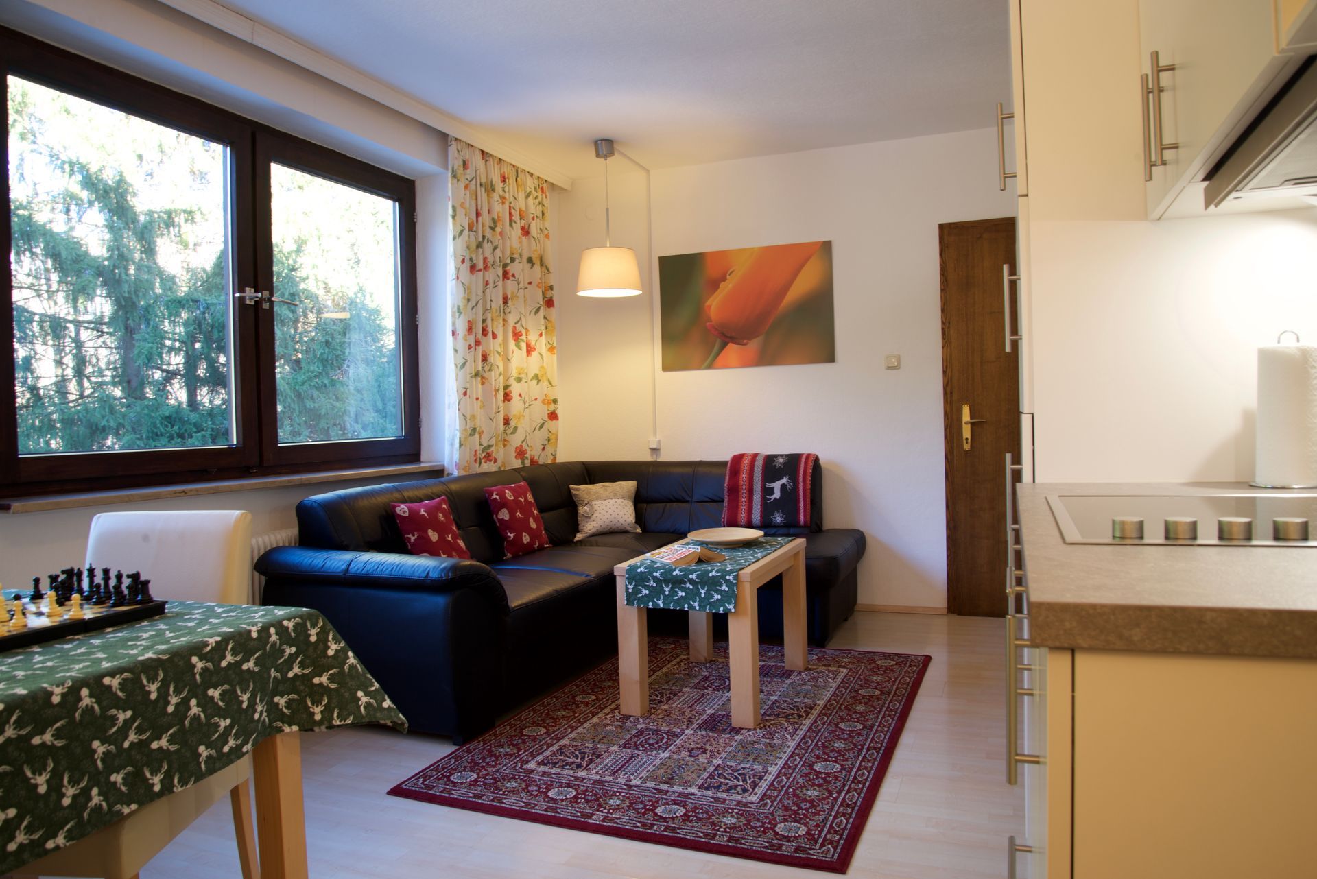 Helles Wohnzimmer mit schwarzem Sofa, rotem Teppich, großem Fenster, farbenfrohen Vorhängen und einem kleinen Tisch.