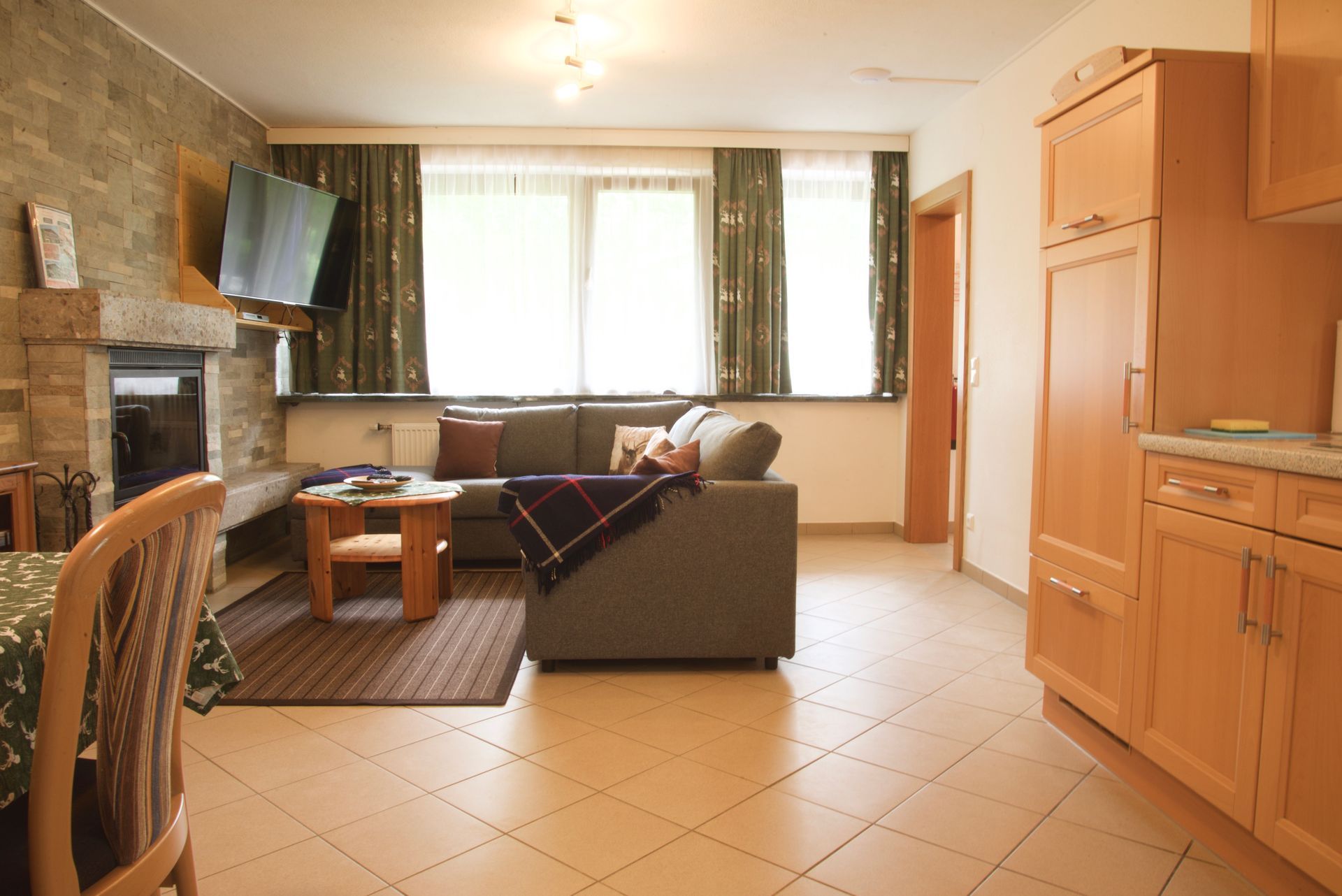 Modernes Wohnzimmer mit grauem Sofa, an der Wand montiertem Fernseher, Holzschränken und hellen Fenstern