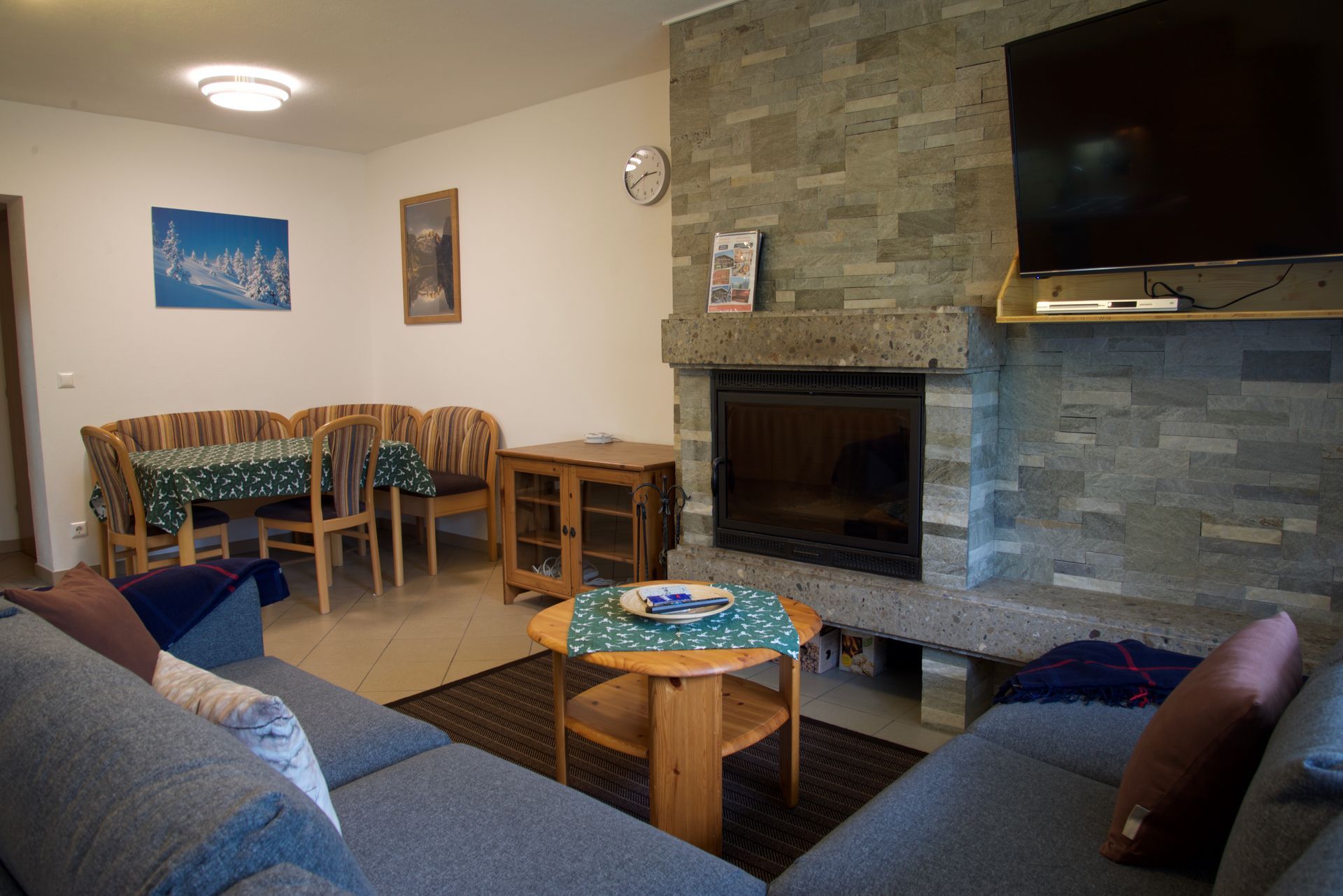 Gemütliches Wohnzimmer mit grauen Sofas, Holztisch, Steinkamin, Wandfernseher und Essbereich im hinteren Bereich