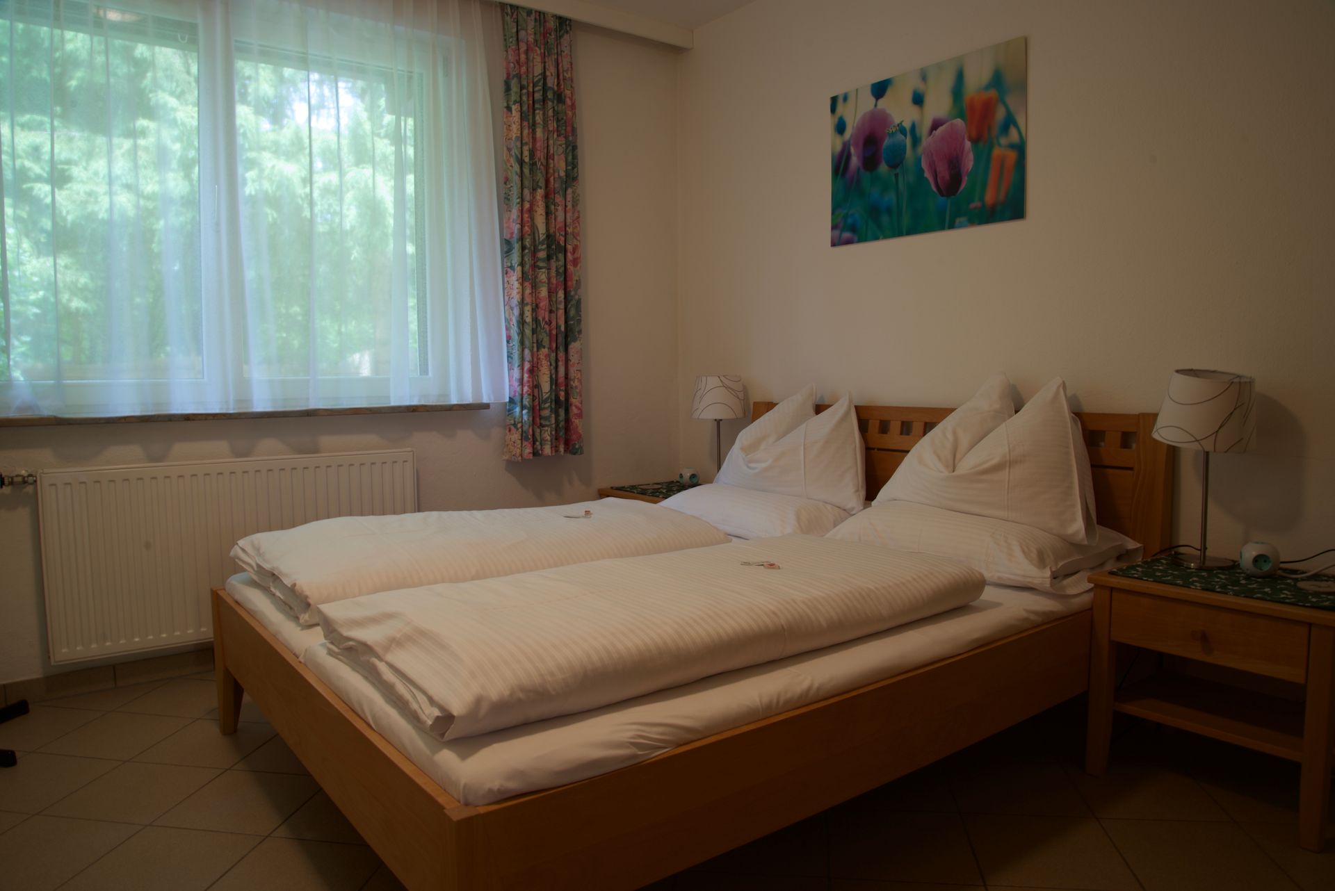 Schlafzimmer mit Doppelbett, weißer Bettwäsche, Nachttischlampen und einem Fenster mit transparenten Vorhängen.