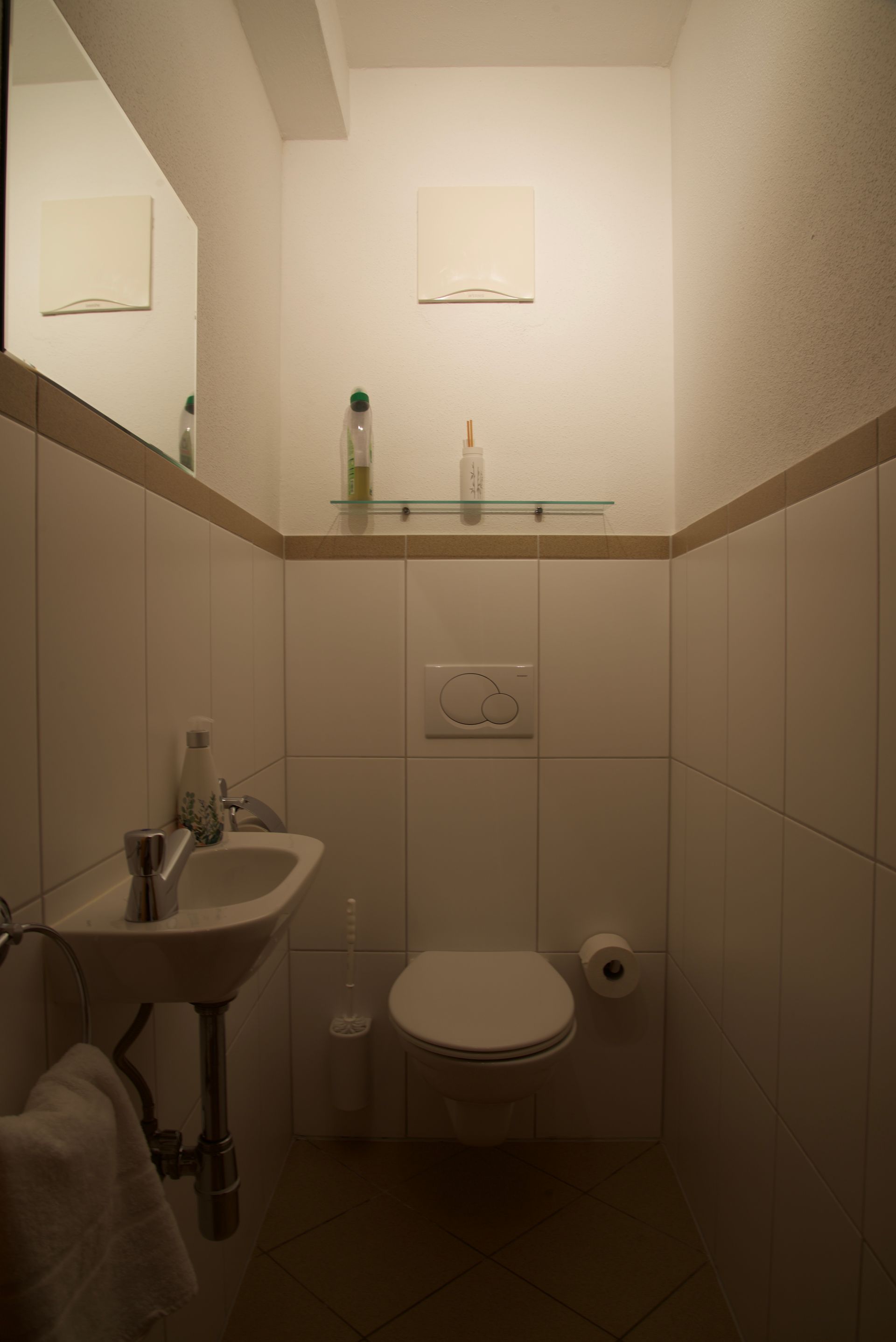 Kleine Toilette mit wandmontiertem WC, Waschbecken, Spiegel und Ablage über gefliesten Wänden.