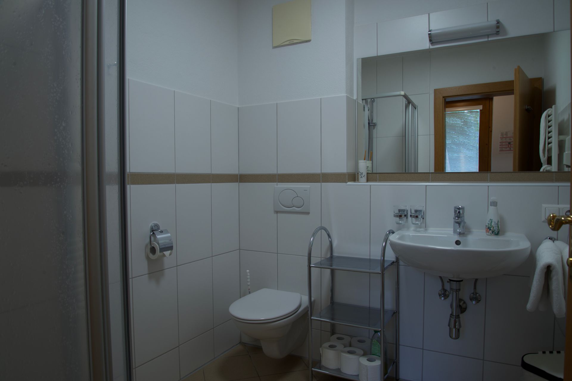 Kleines gefliestes Badezimmer mit Toilette, Waschbecken, Spiegelschrank und einem Metallrollator neben dem Waschbecken.
