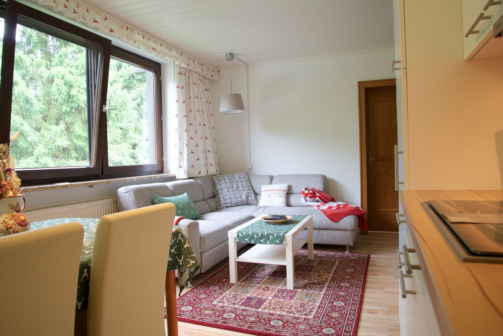 Helles Wohnzimmer mit grauer Eckcouch, rotem Teppich, Couchtisch, Esszimmerstühlen und großem Fenster mit Blick ins Grüne