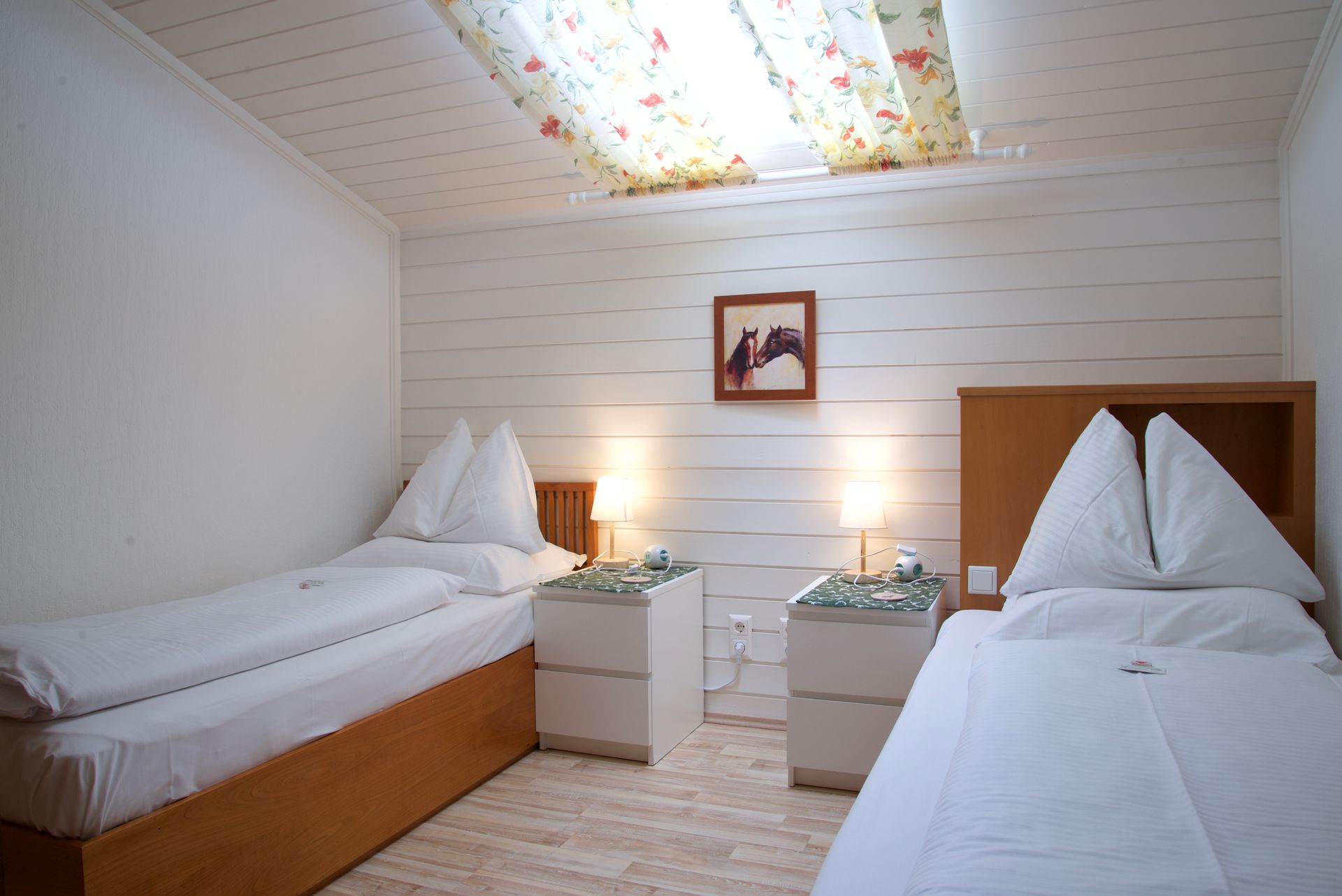 Kleines Hotelzimmer mit zwei Einzelbetten, Nachttischlampen, weiß getäfelten Wänden und einem farbenfrohen Dachfenster.