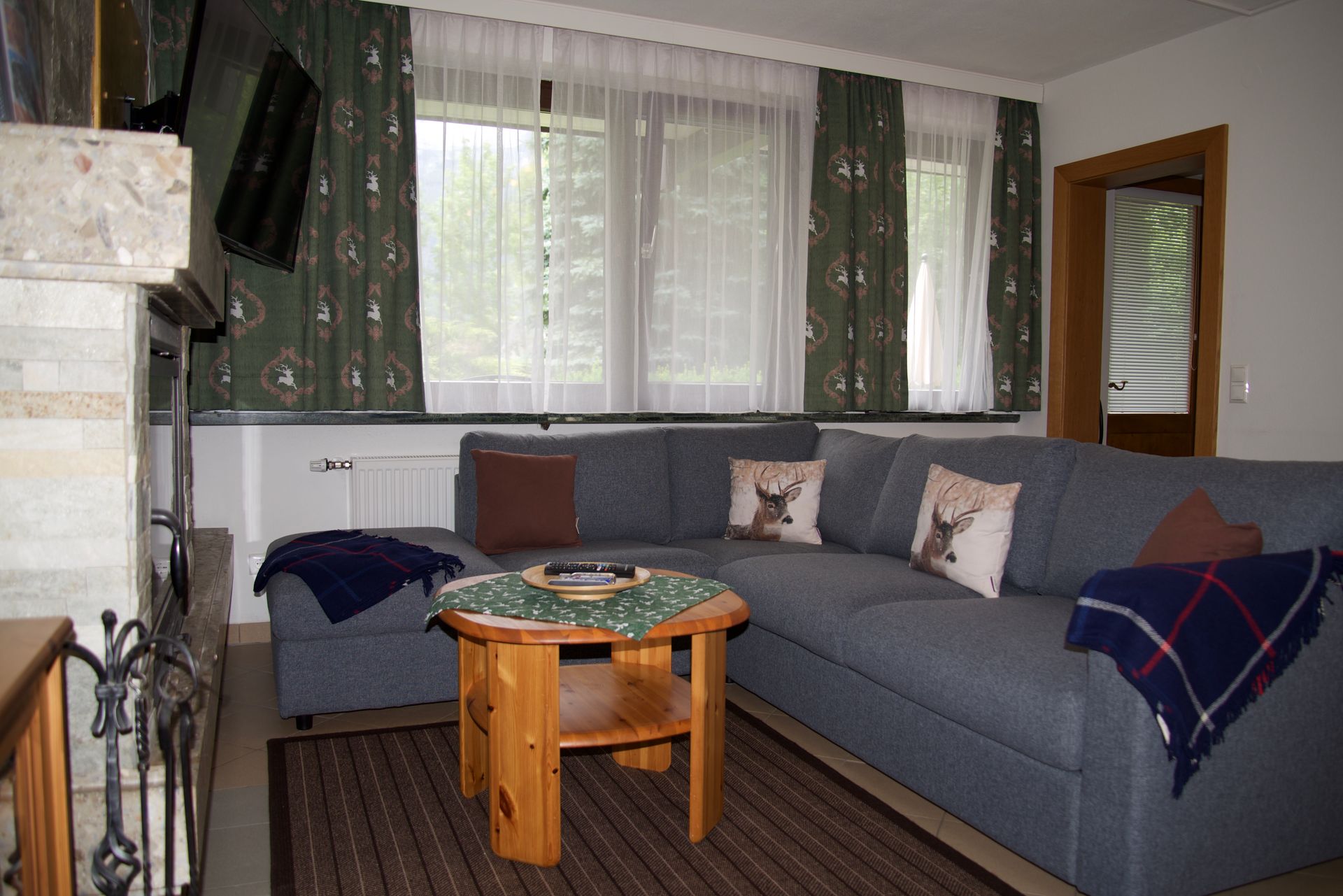 Gemütliches Wohnzimmer mit grauem Ecksofa, Couchtisch aus Holz, Kamin und großem Fenster mit Vorhängen