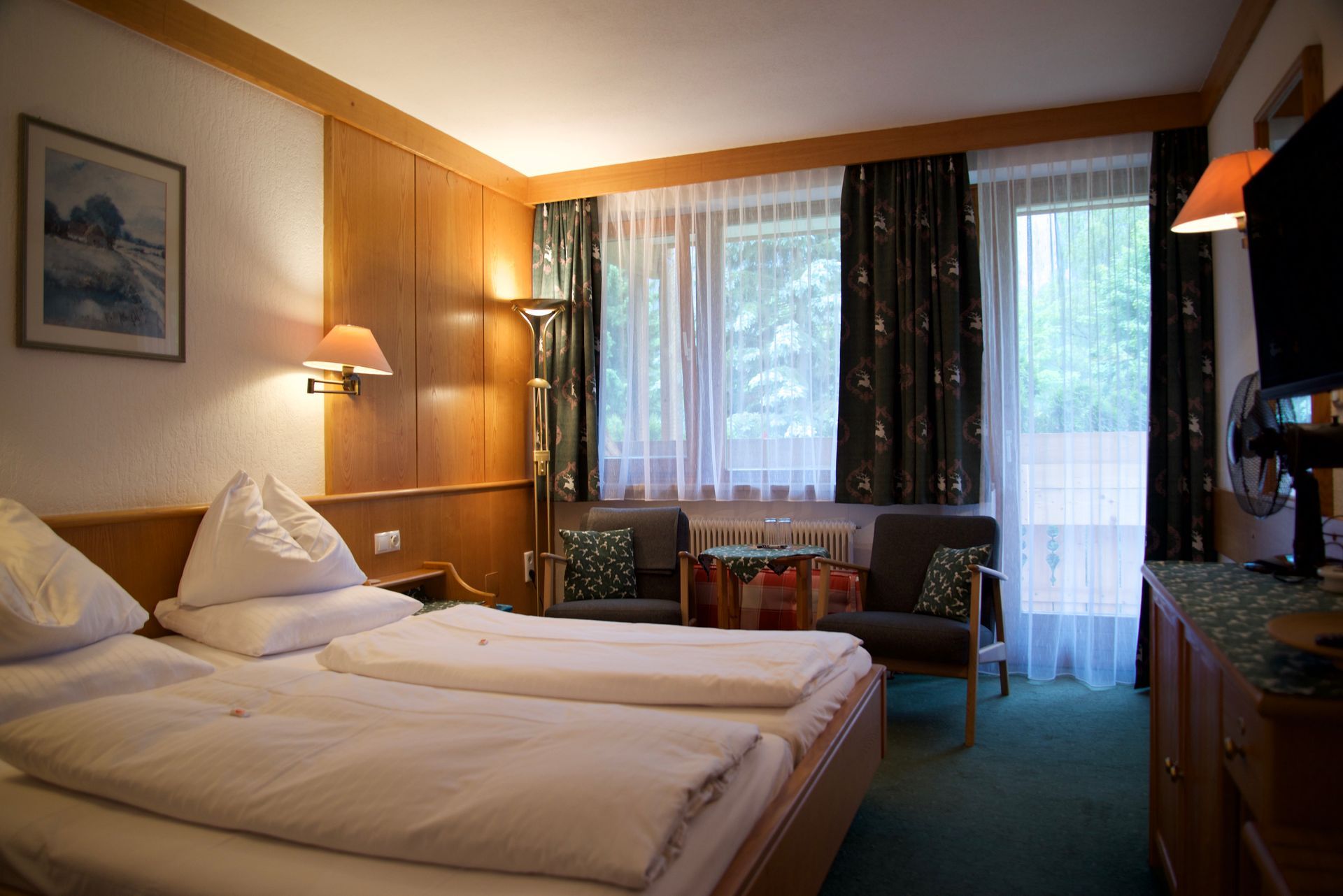 Hotelzimmer mit zwei Einzelbetten, warmer Beleuchtung und einem Fenster mit Vorhängen mit Blick auf Bäume.