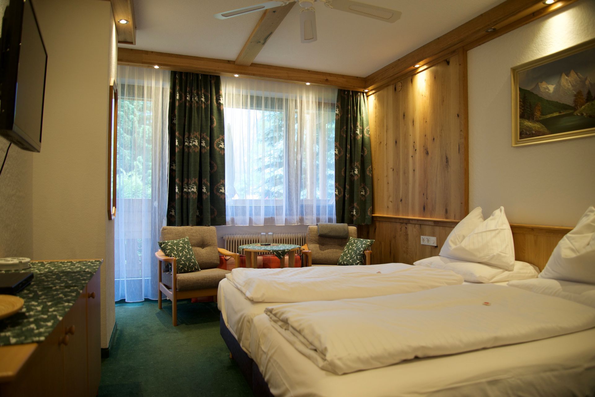 Hotelzimmer mit zwei Betten, großem Fenster mit Vorhängen, Sitzecke und warmer Holzvertäfelung