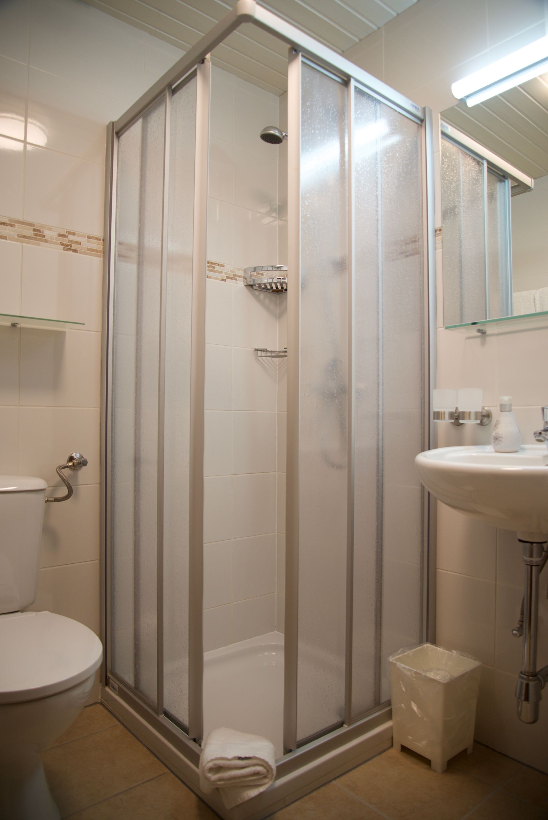 Kleines Badezimmer mit Duschkabine aus Glas, Toilette, Waschbecken, Spiegel und beige gefliesten Wänden.