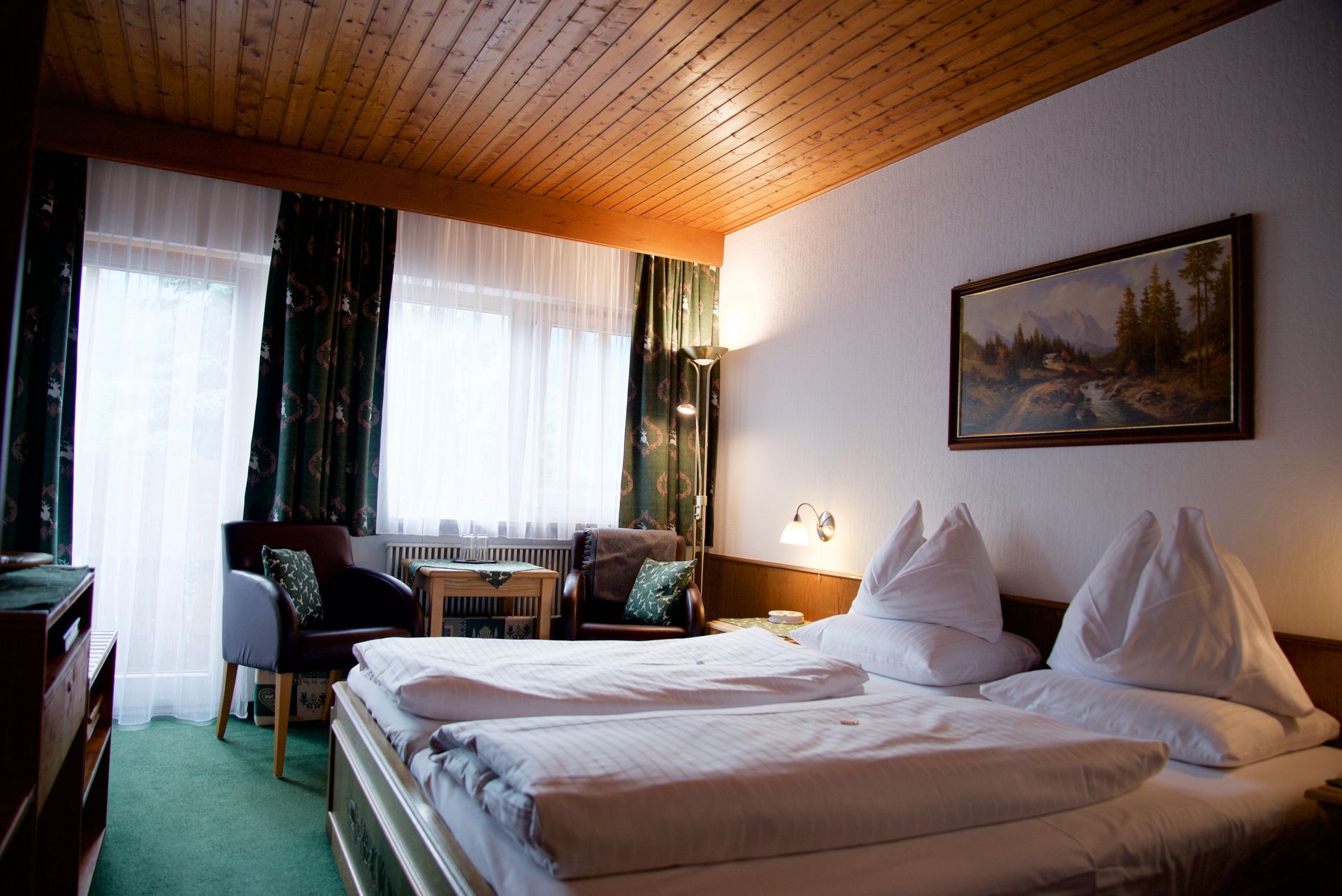 Hotelzimmer mit zwei Einzelbetten, warmer Holzdecke, Vorhängen und einer Sitzecke am Fenster.