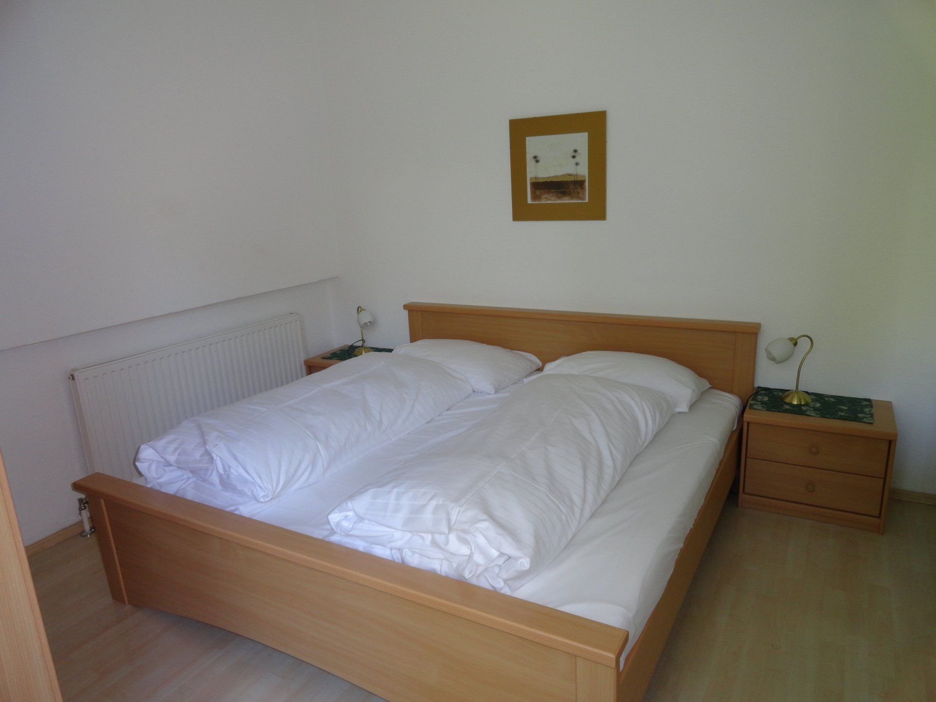 Kleines Schlafzimmer mit Holzbett, weißer Bettwäsche, Heizkörper, Nachttisch und Wandspiegel