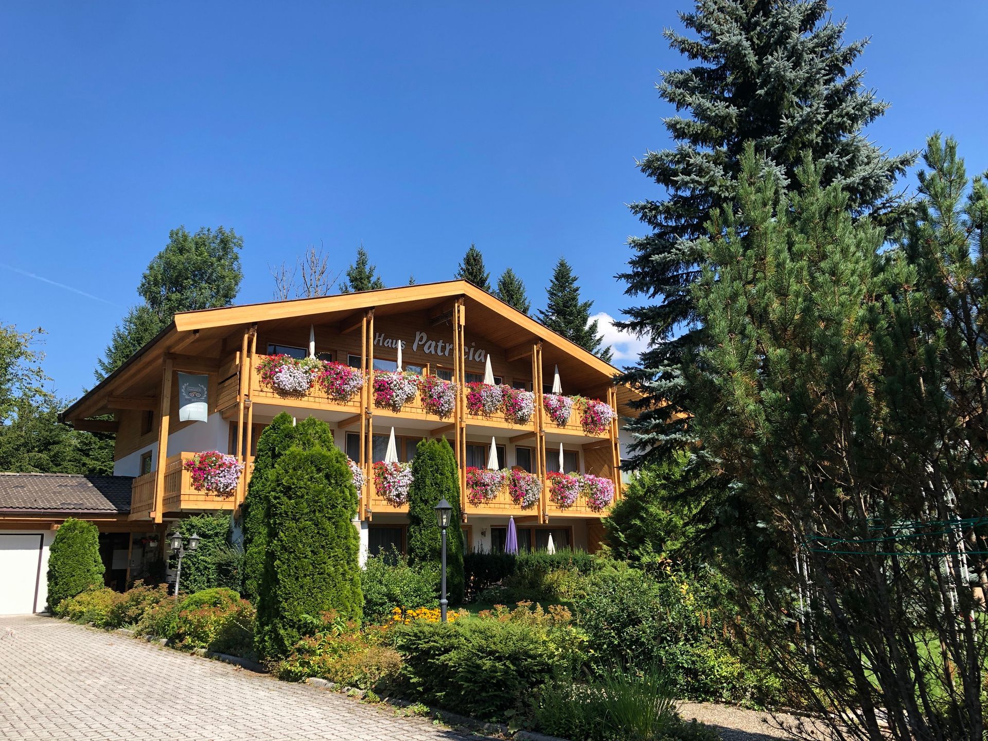 Ein hölzernes Chalet mit blumengeschmückten Balkonen neben einer Auffahrt unter strahlend blauem Himmel