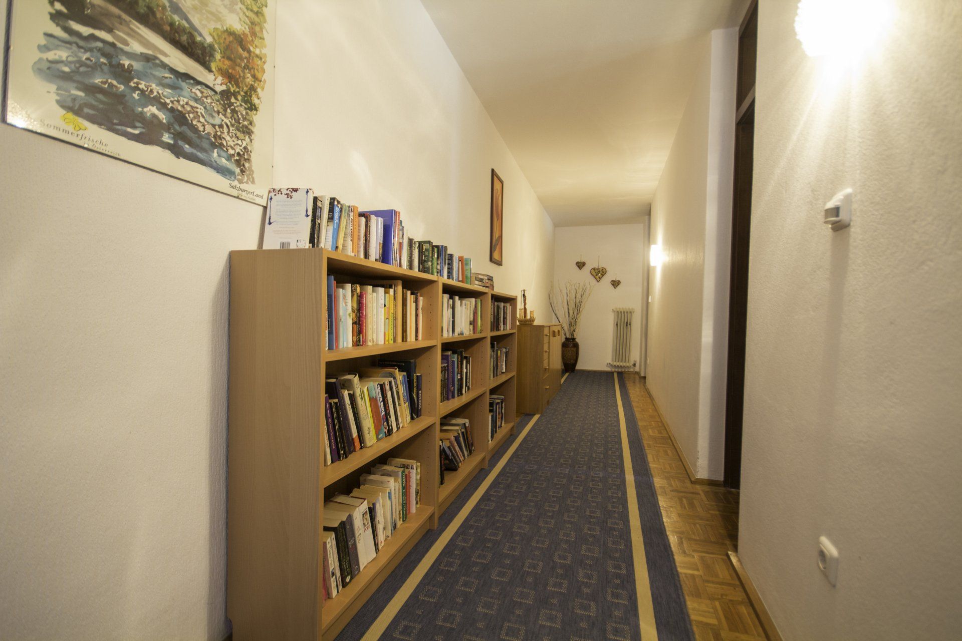 Schmaler Flur mit Bücherregalen auf der linken Seite, blauem Teppichläufer und warmen Wandleuchten.