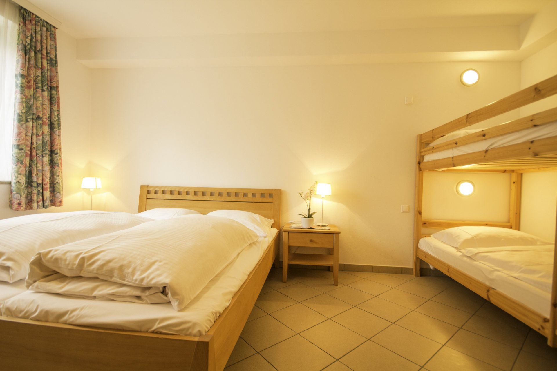 Helles Hotelzimmer mit Doppelbett, Etagenbett, Nachttischen und warmem Lampenlicht