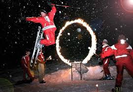 Ein Feuerkünstler in rotem Overall springt nachts durch einen brennenden Reifen, Helfer sind in der Nähe.