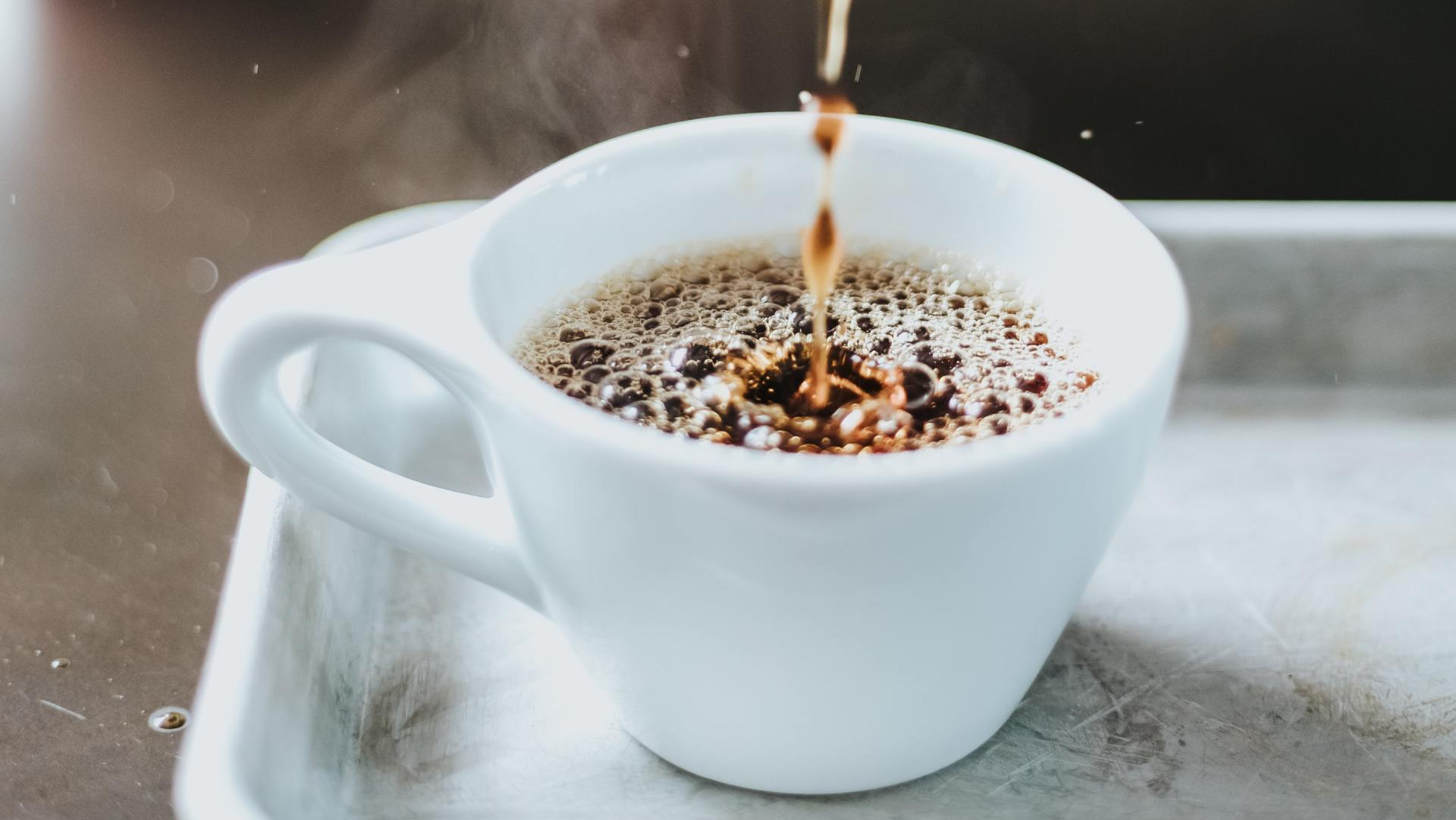 Kaffee wird in eine weiße Tasse gegossen, Dampf steigt von einer Arbeitsplatte auf.