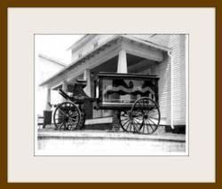 Black-and-white photo of a horse-drawn wagon parked beside a building under a porch roof