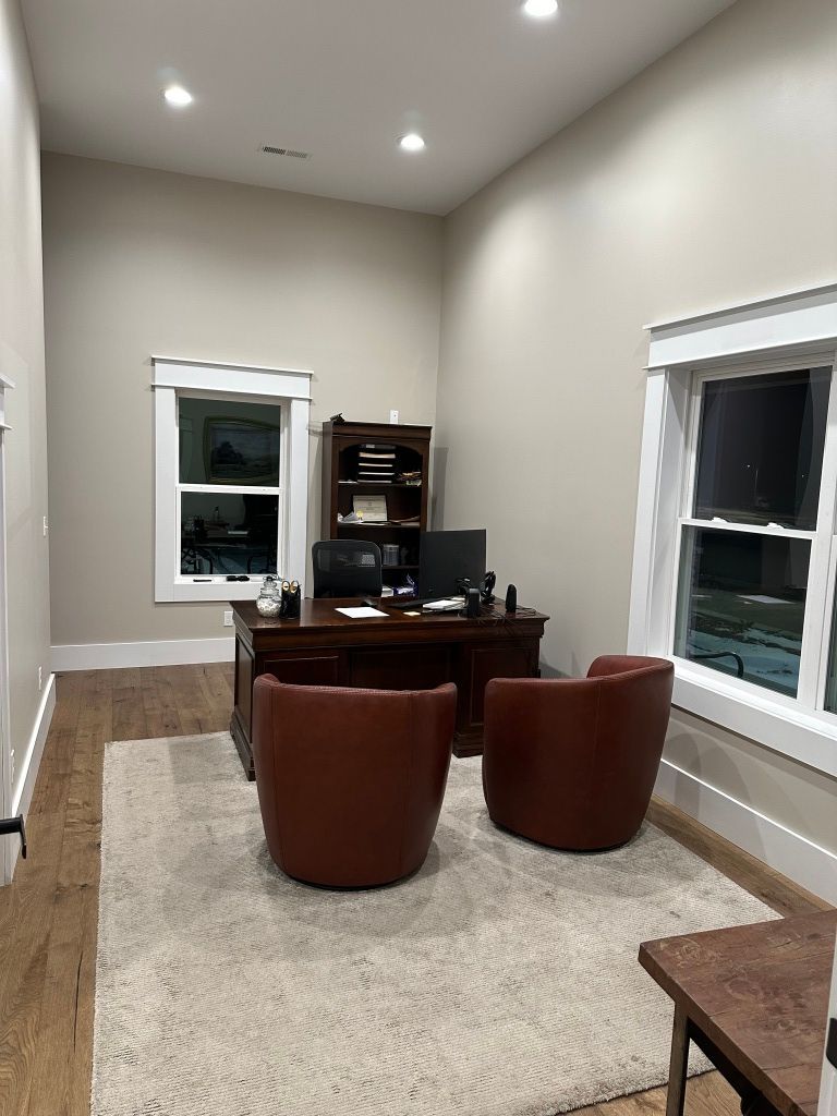 Small office with desk, computer, two brown chairs, cream rug, and windows on both sides.