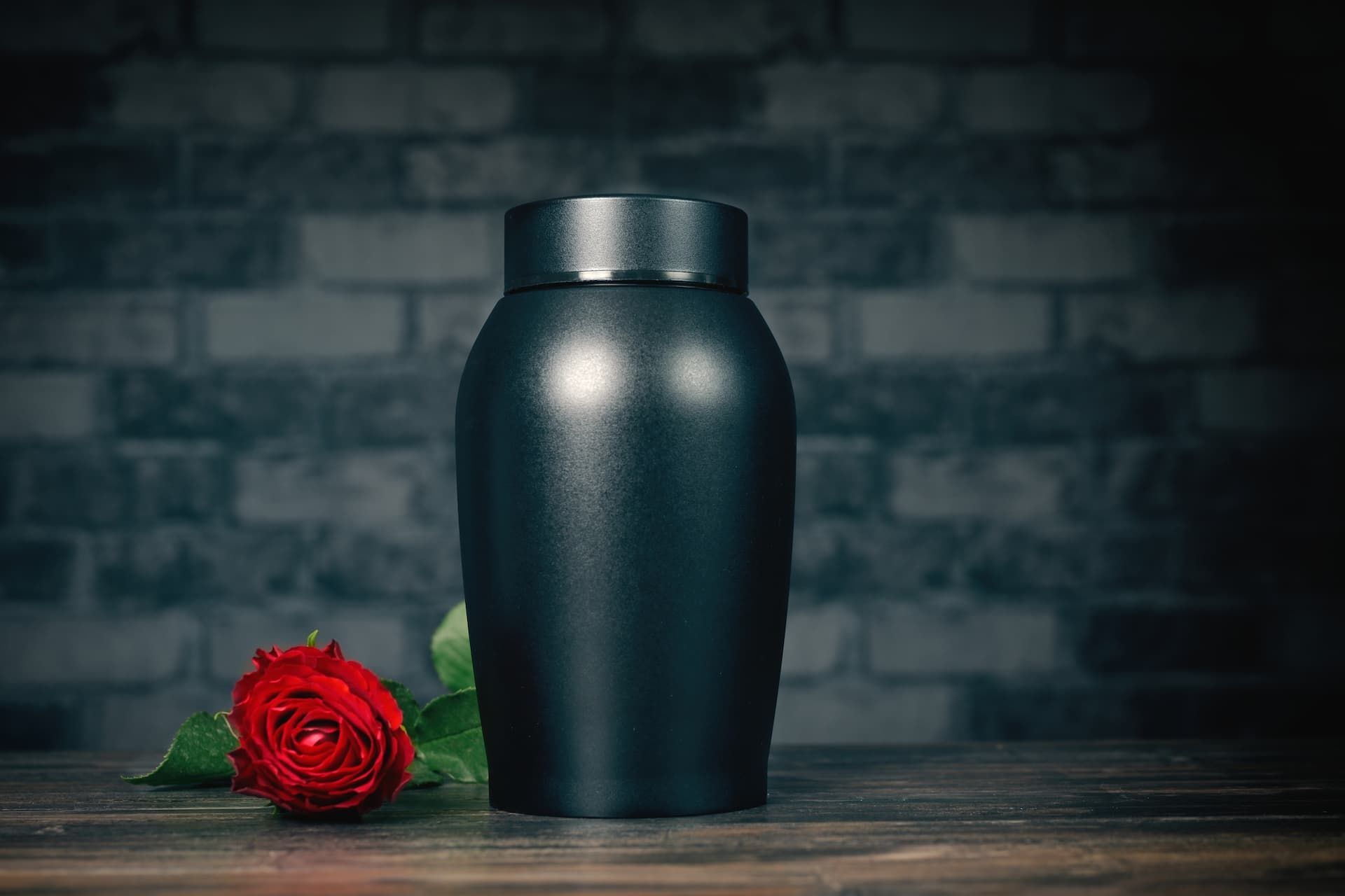 Black urn on a wooden surface with a red rose beside it against a dark brick wall.