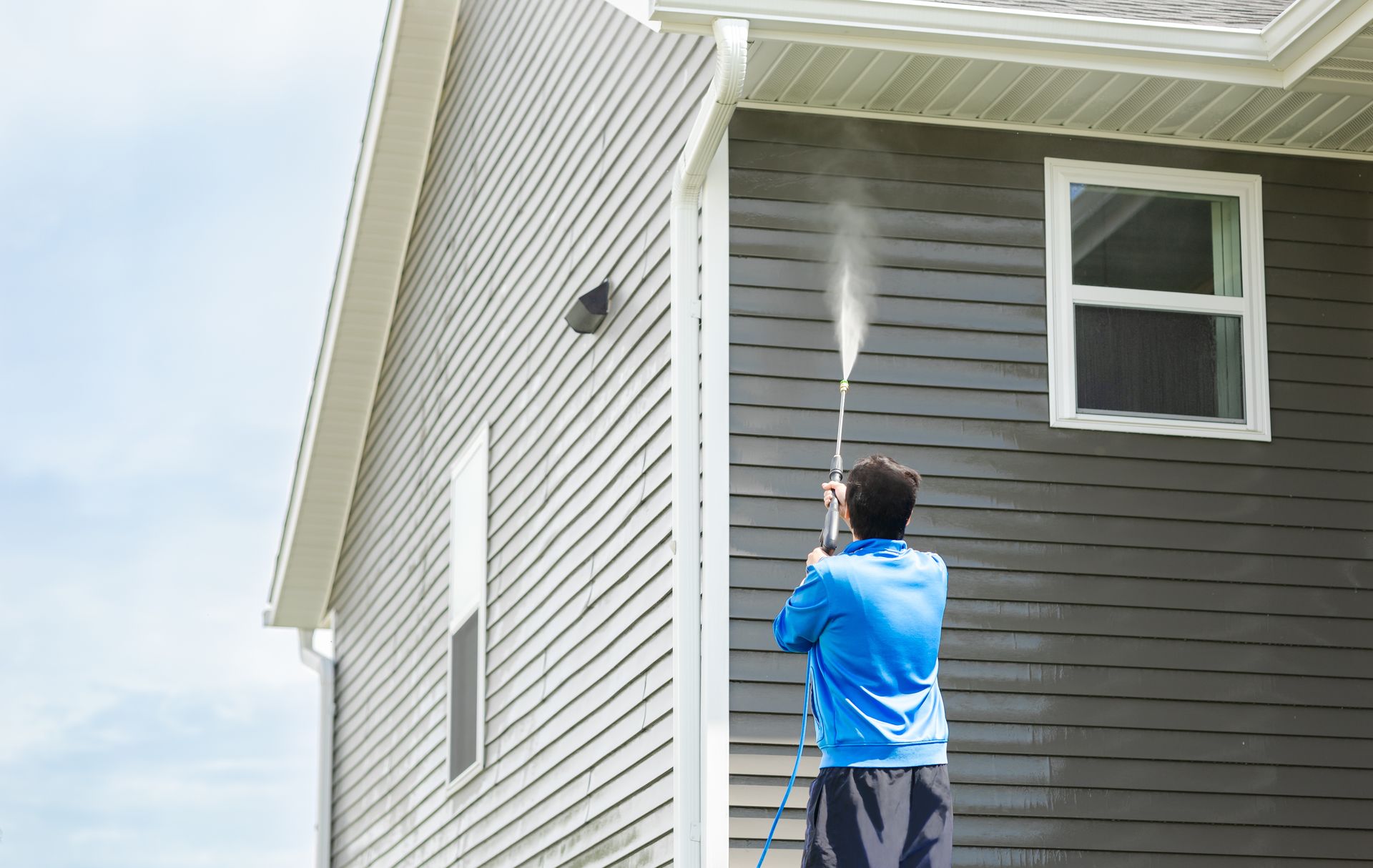 A person in a blue jacket pressure washes the dark gray siding of a two-story house.