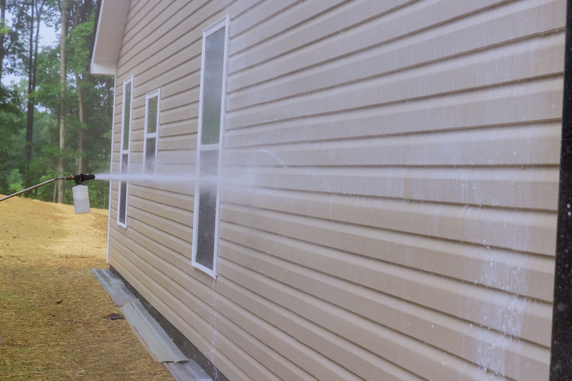 A pressure washer sprays cleaning solution onto the beige vinyl siding of a house exterior.