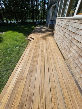 A person in a plaid shirt uses a roller to stain wooden deck planks dark brown outdoors on a sunny day. A person in a plaid shirt uses a roller to stain wooden deck planks dark brown outdoors on a sunny day.