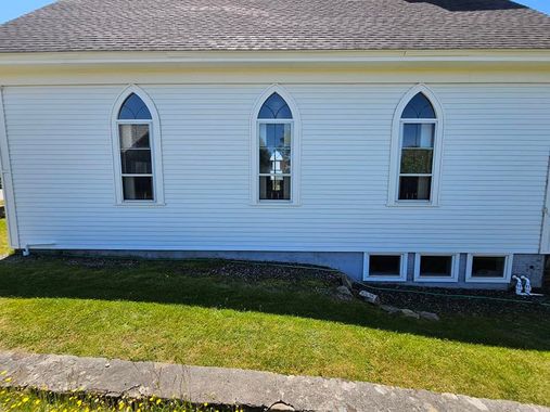 A person in a neon yellow jacket on a ladder uses a pressure washer to clean the side of a white-sided house. A person in a neon yellow jacket on a ladder uses a pressure washer to clean the side of a white-sided house.