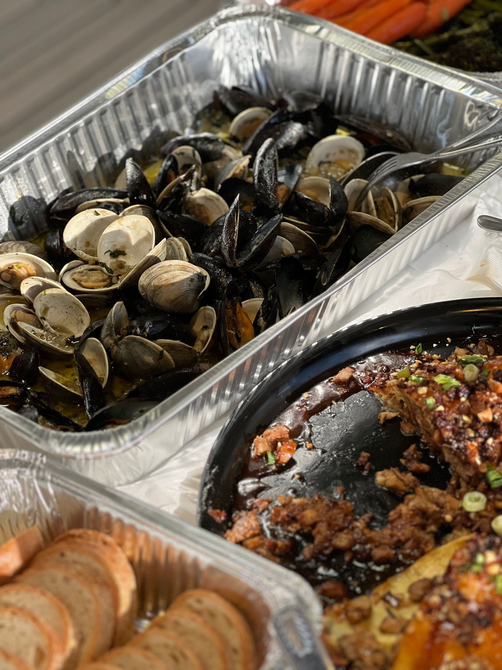 Trays of cooked clams, saucy seafood, and sliced bread at a buffet.