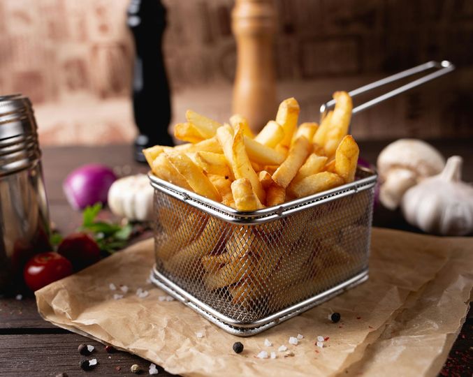 Fried French fries in a metal mesh basket on a rustic wooden board, with garlic and peppers in the background