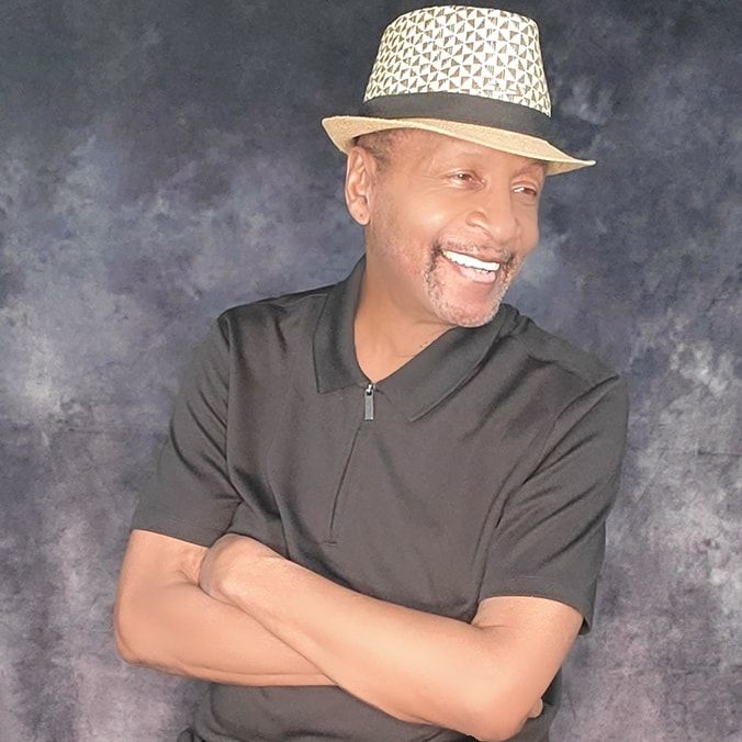 A man wearing a hat and a black shirt is smiling with his arms crossed.