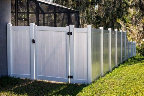 White vinyl fence with gate along a grassy yard, beside a black chain-link fence and trees