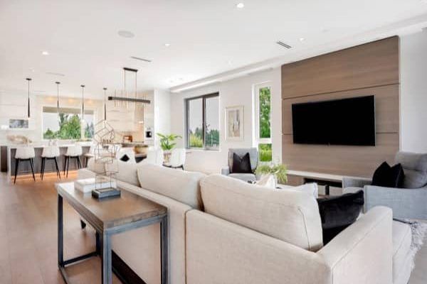 Bright open-plan living room with beige sectional, wall-mounted TV, and dining area in the background