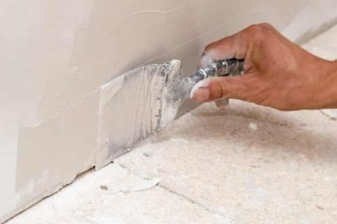 Hand spreading white compound along a wall baseboard with a putty knife