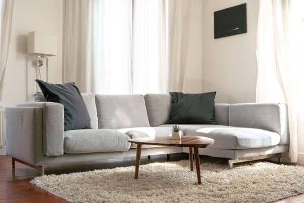 Bright living room with a gray sectional sofa, round wooden coffee table, and light curtains.