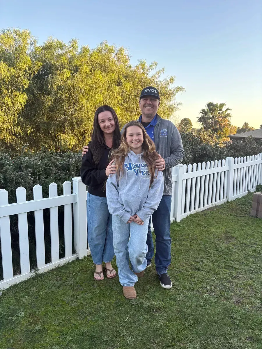 Family of three smiling, posing by a white picket fence on a sunny day.