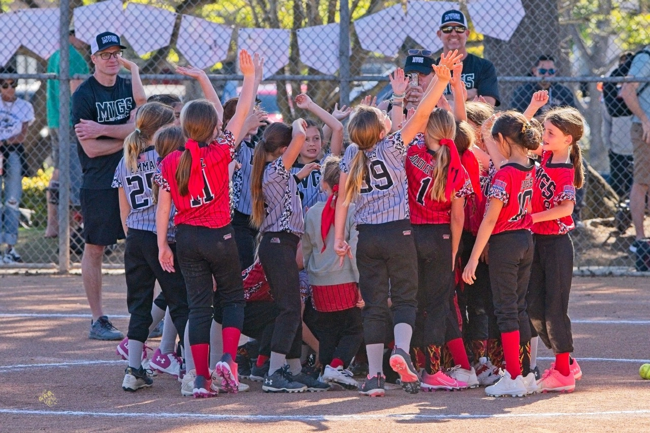 Youth softball team huddles on the field, arms raised in a group cheer.