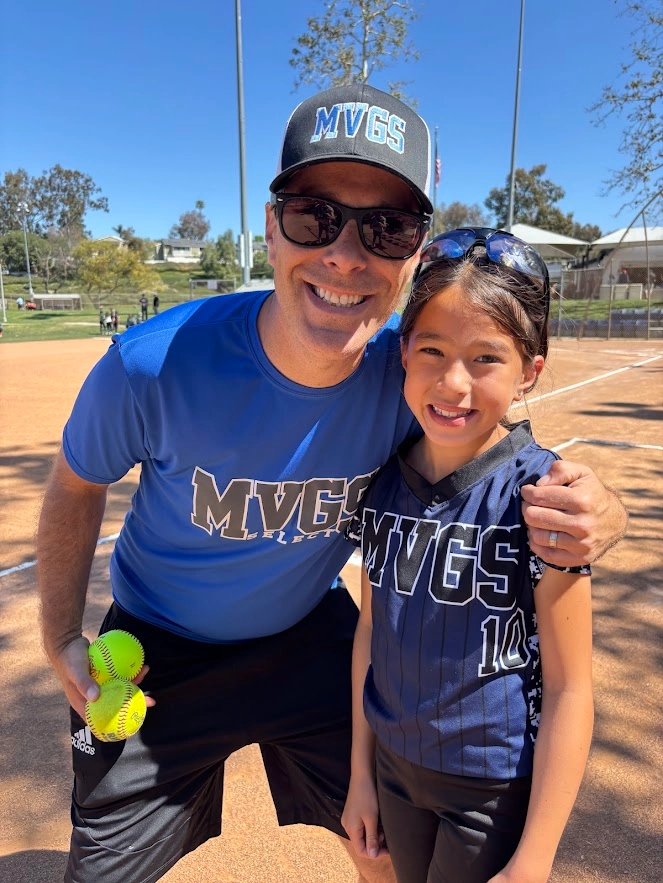 Man in blue shirt and baseball cap with a young girl in a softball uniform on a field, smiling.