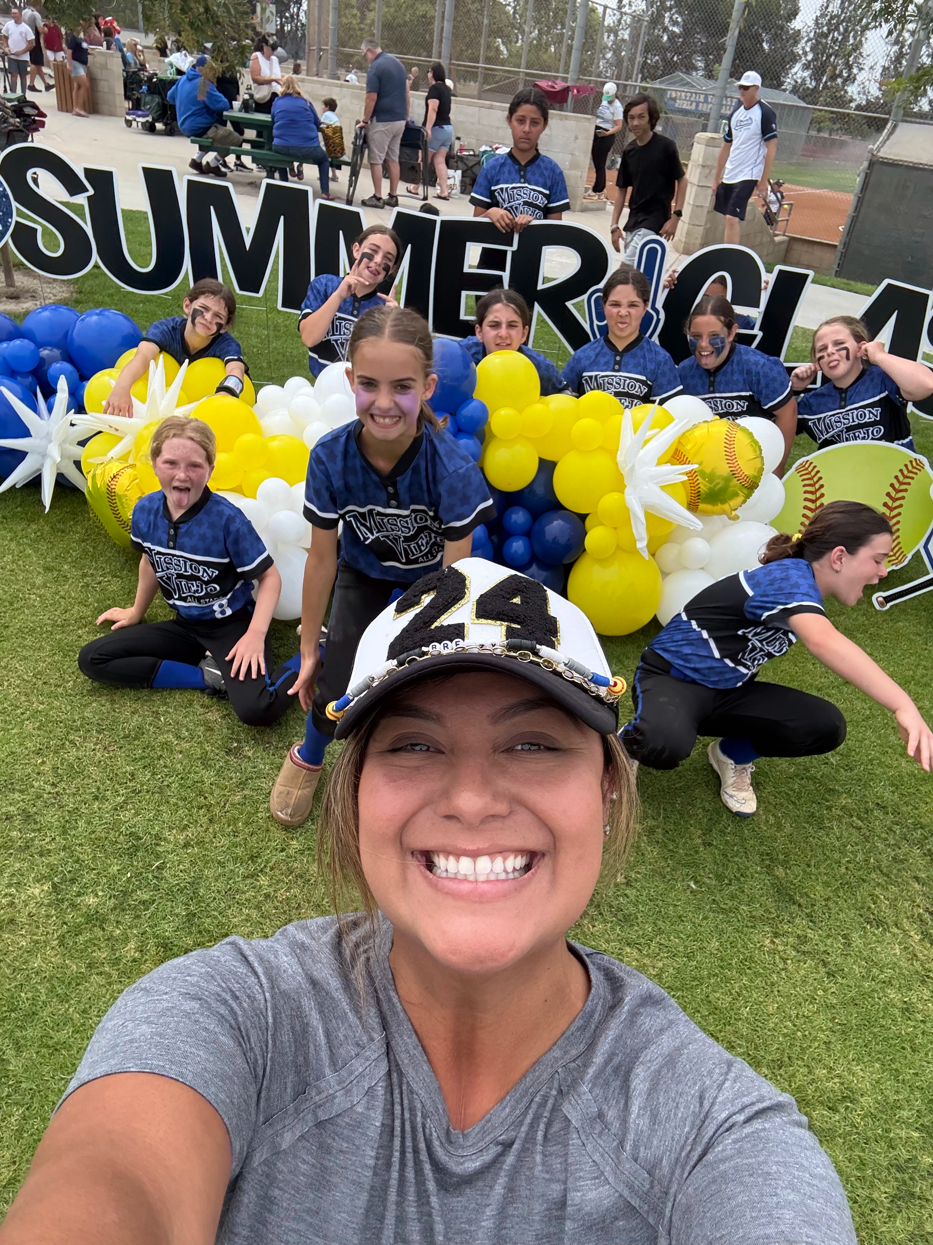Softball team with blue and yellow balloons poses on grass, holding signs. Players smile in the summer sun.