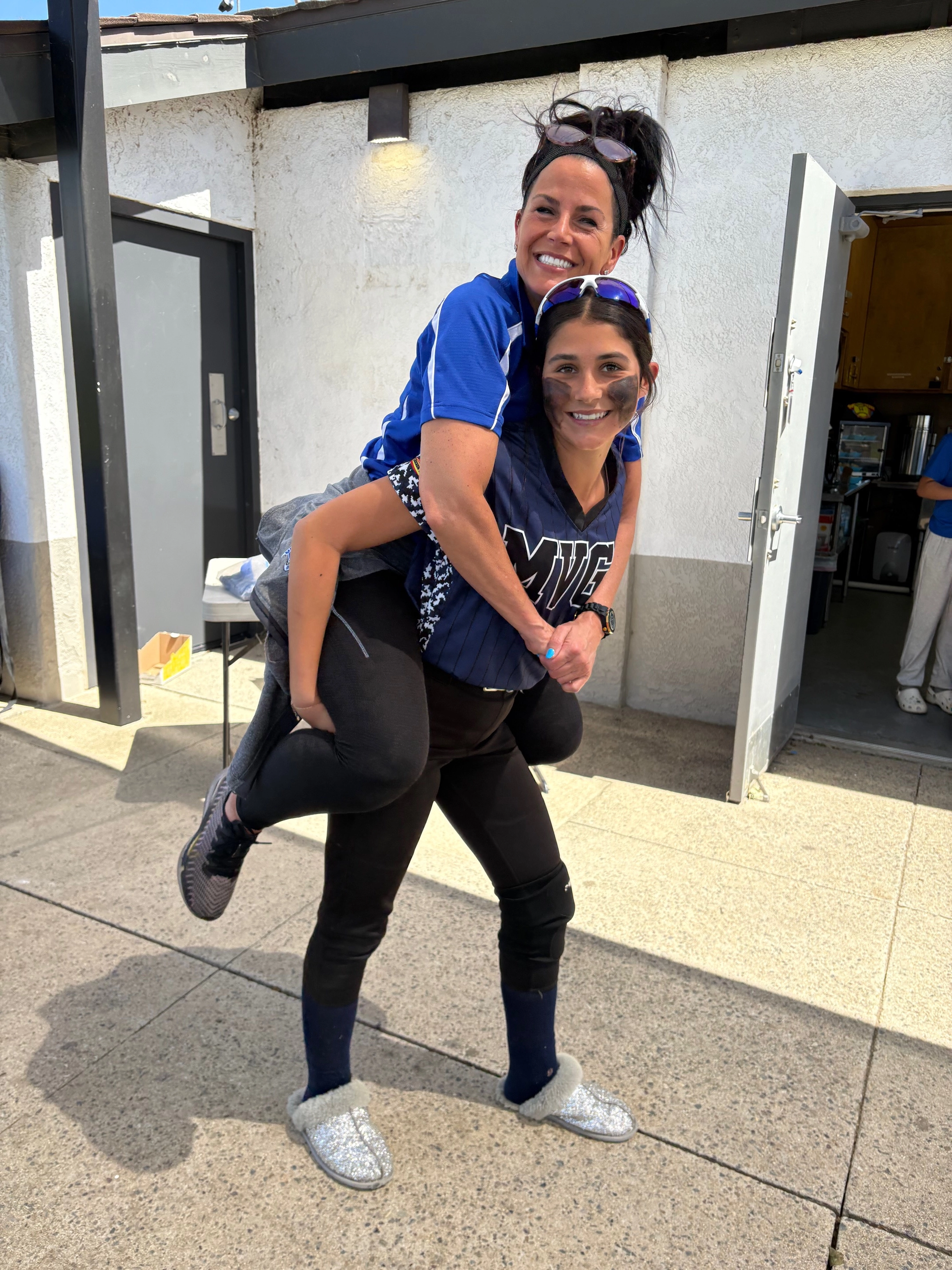 Woman giving a piggyback ride to another woman, both smiling outdoors.