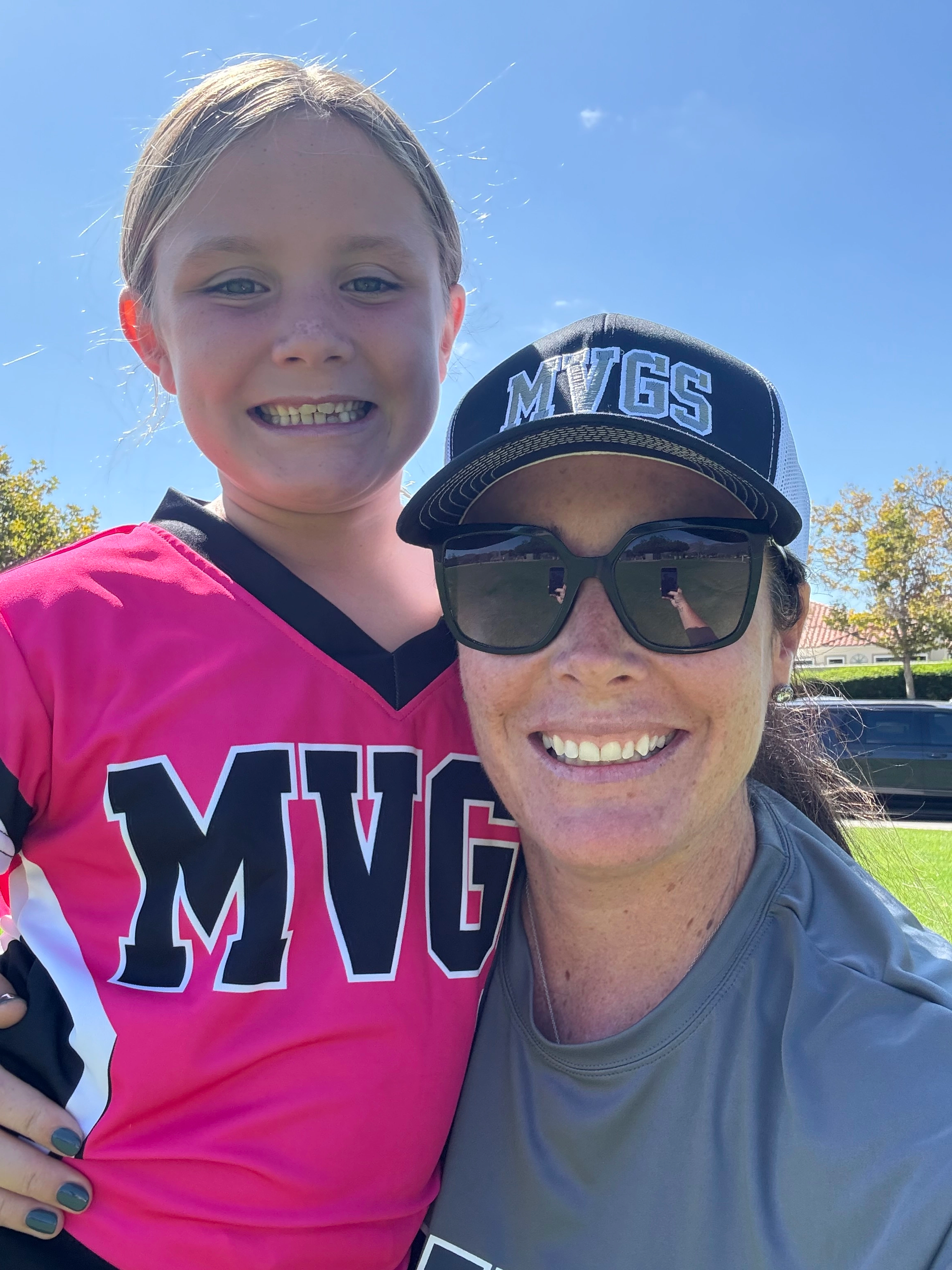 Woman in sunglasses and a girl in a pink jersey smile outdoors. Sunny day.