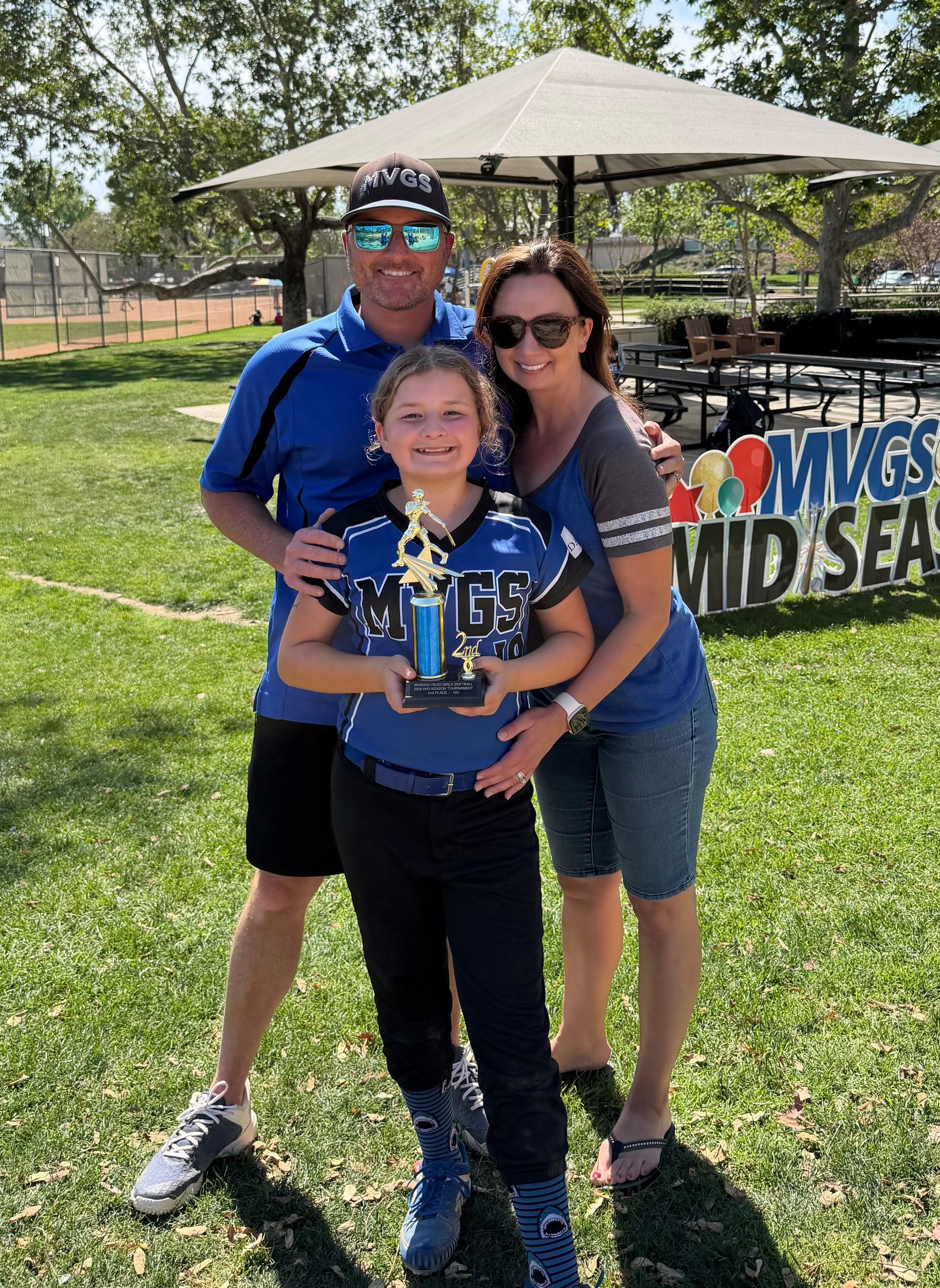 Three people posing outdoors, holding a trophy in front of a grassy field and canopy tent.