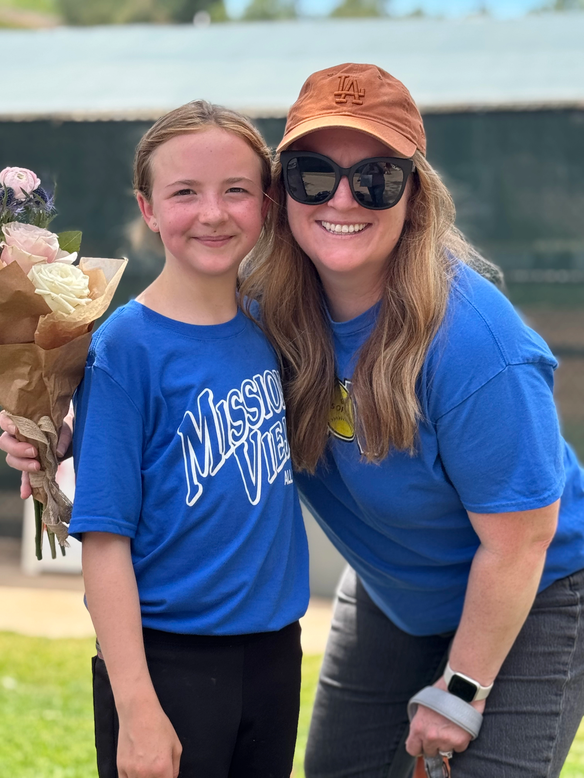 Woman in cap with girl holding flowers, both wearing blue shirts, smiling outside.