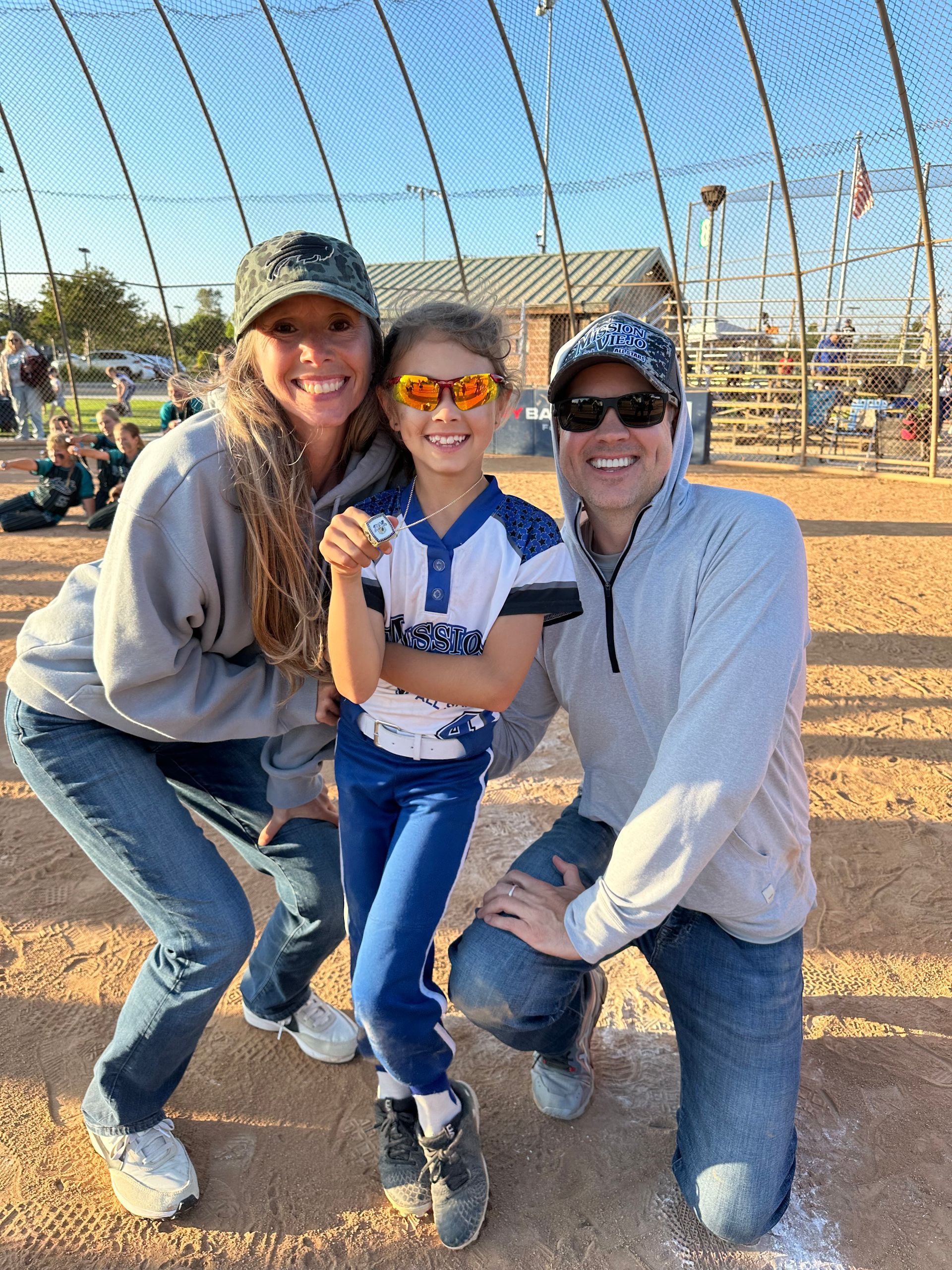 Three people smiling and posing outdoors at a sunny baseball field behind a netting fence.