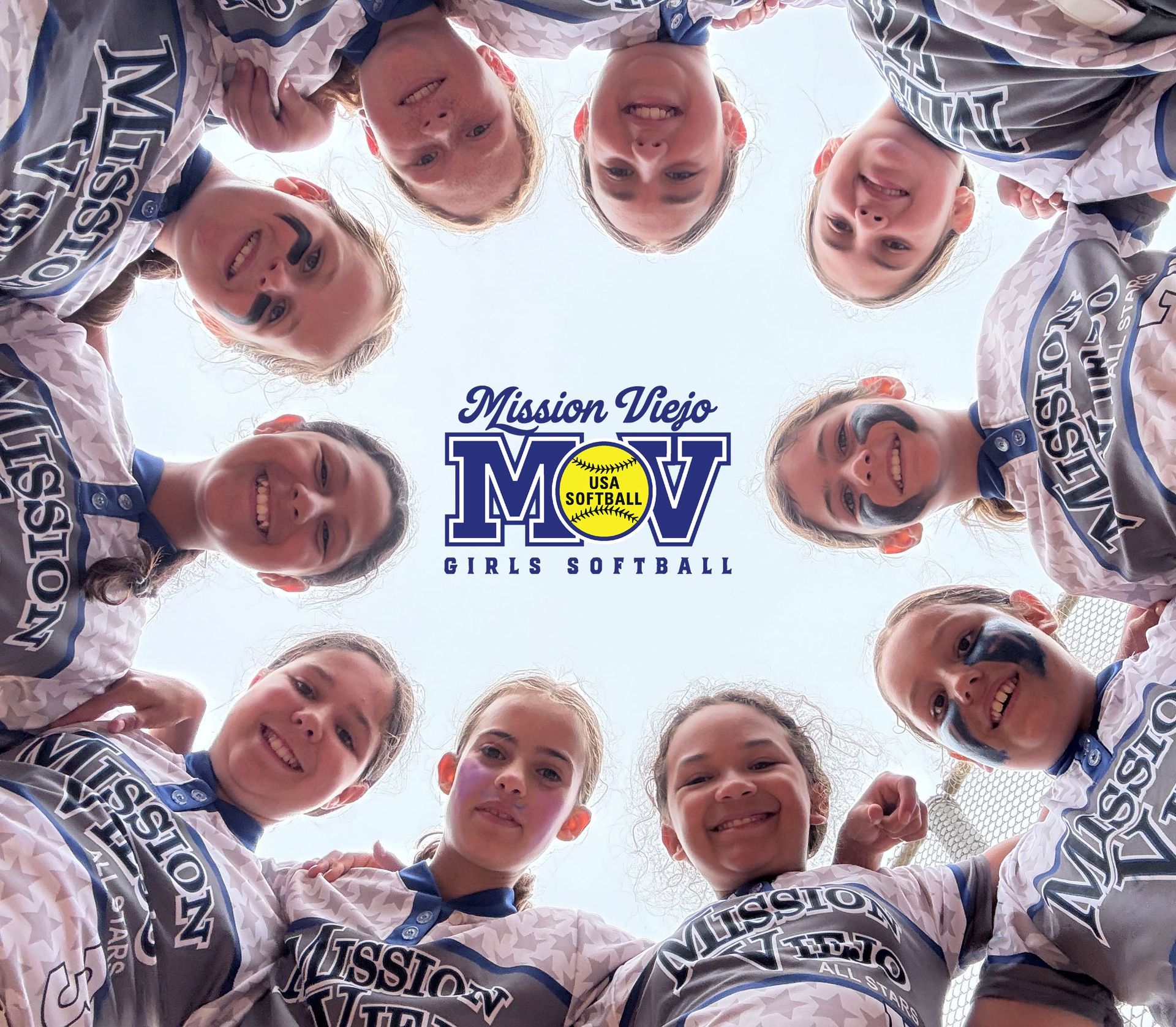 Girls softball team huddles, looking up, arms around each other.
