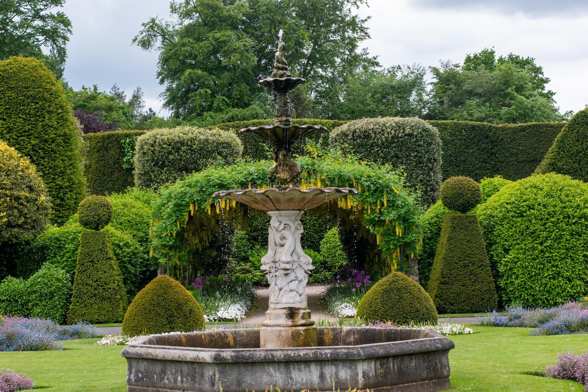 Stone fountain in a formal garden with manicured hedges and flowering plants.