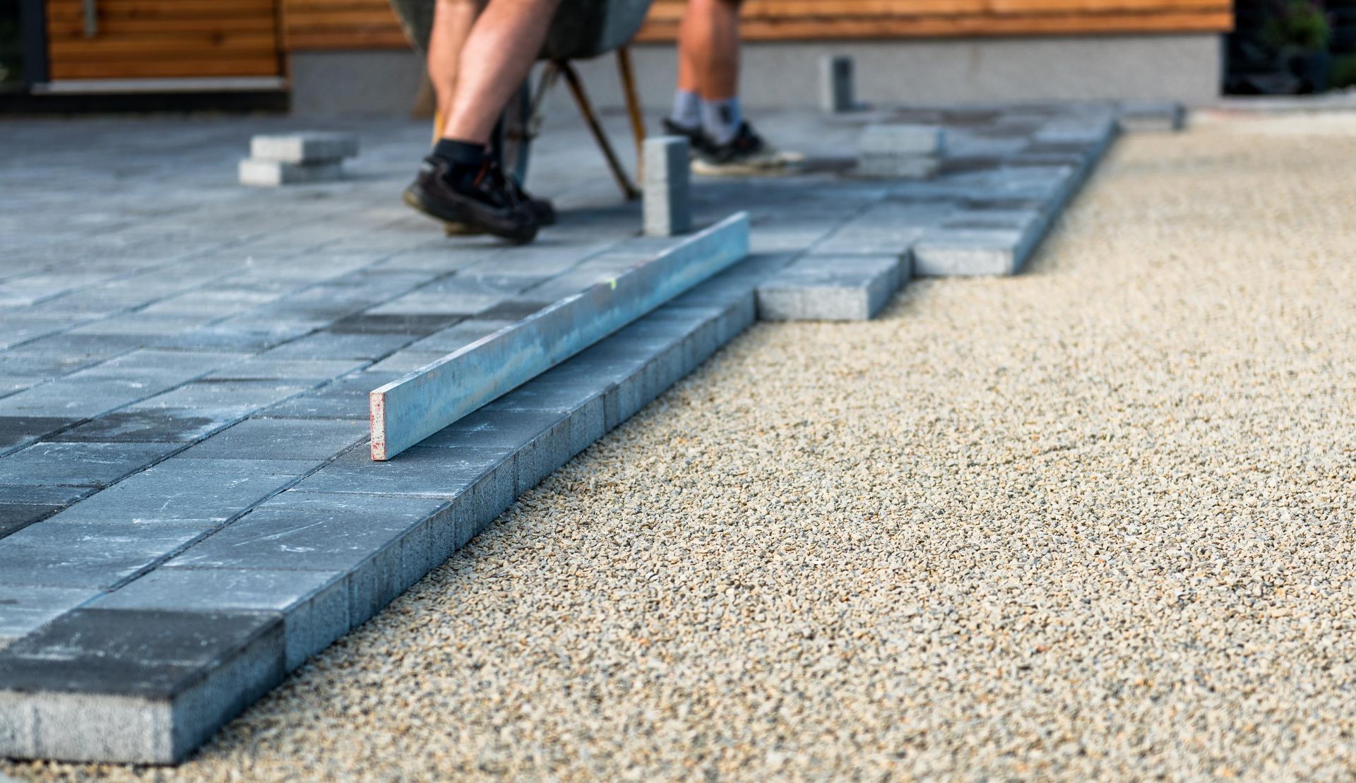 Workers laying paving stones, using a level, on a gravel base. Blue-gray stones, daylight.