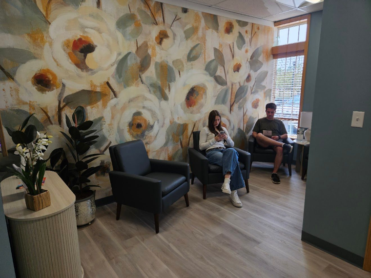 Waiting area with floral mural, two people seated on chairs, and a large potted plant.