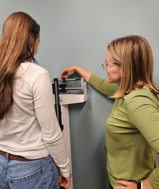 Two people measuring a woman’s weight on a scale against a gray wall