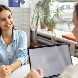 Two people in a bright office during a meeting, with one smiling across a laptop